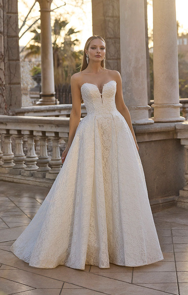Woman in 63110 Huntley by Veni Infantino - Strapless Lace Ball Gown with Low Sweetheart Neckline standing in an elegant outdoor setting with columns and sunlight.