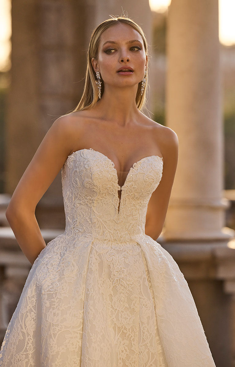 Woman wearing 63110 Huntley by Veni Infantino - Strapless Lace Ball Gown with Low Sweetheart Neckline in an outdoor setting with columns.