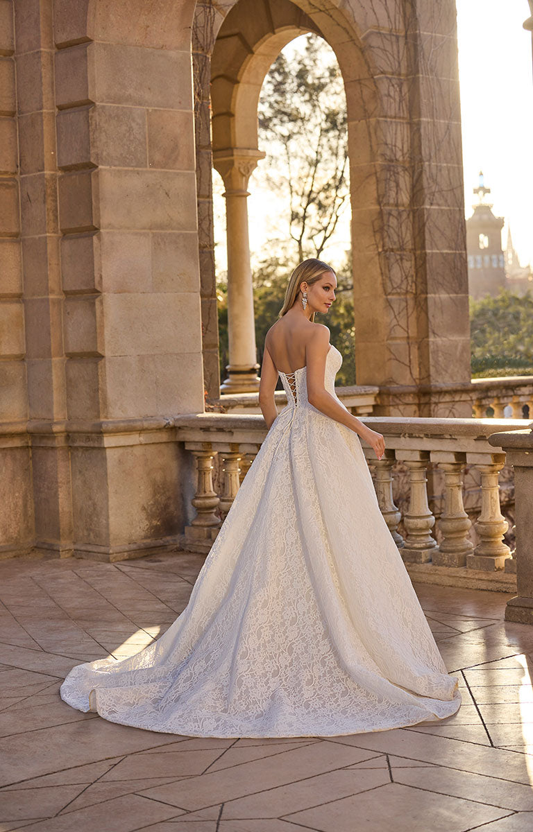 Woman in 63110 Huntley by Veni Infantino - Strapless Lace Ball Gown with Low Sweetheart Neckline standing on a stone balcony with architectural background