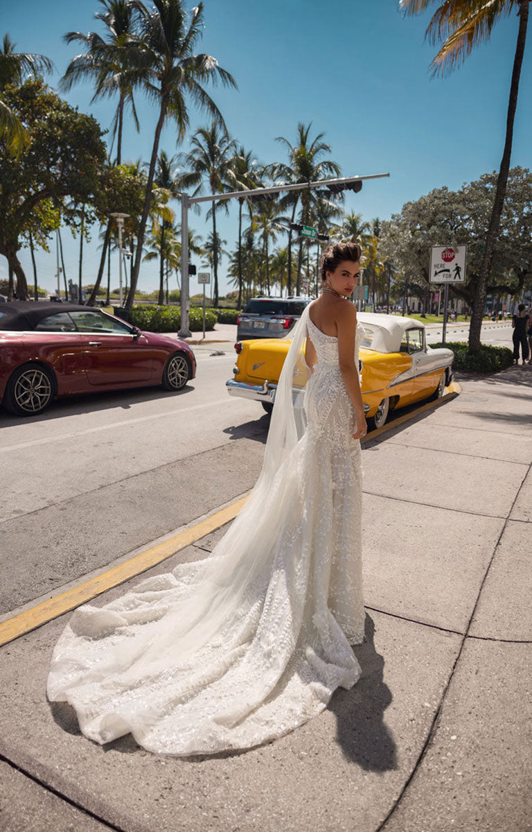 Woman in a white wedding dress standing on a street with palm trees and vintage cars in the background.