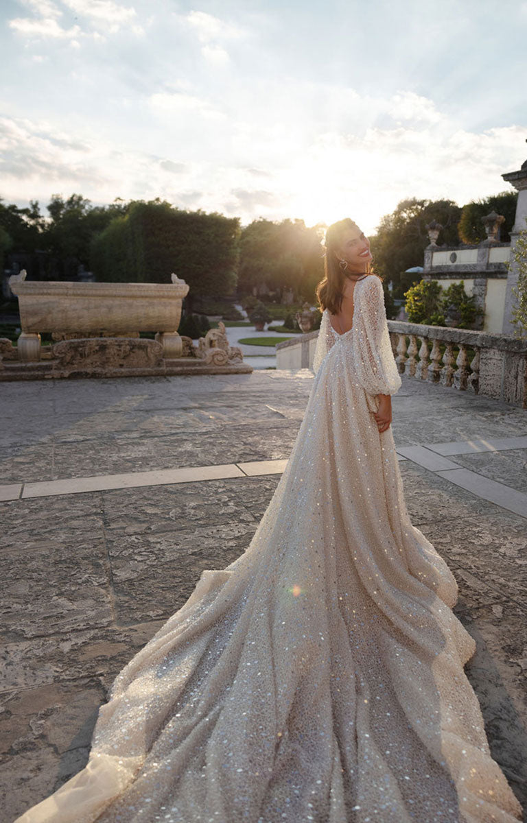Woman in a sparkling wedding dress standing in an outdoor setting with stone pavement and greenery.
